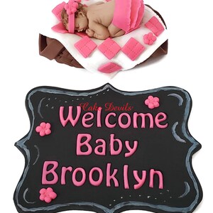 May include: A black chalkboard sign with a white border and pink lettering that reads "Welcome Baby Brooklyn". There are pink flowers on the sign.