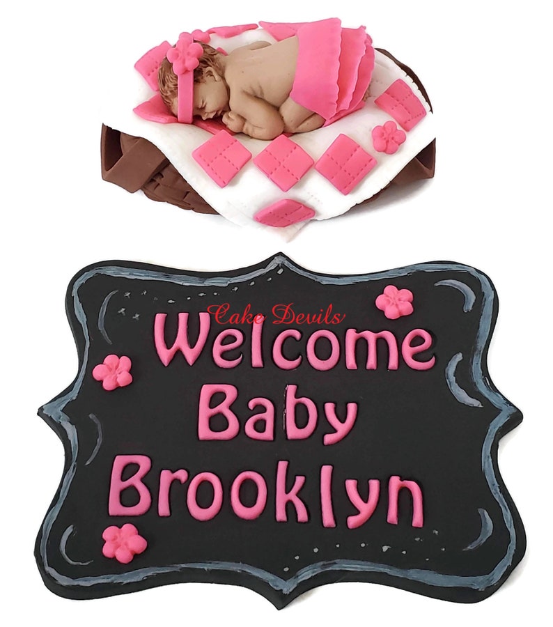 May include: A black chalkboard sign with a white border and pink lettering that reads "Welcome Baby Brooklyn". There are pink flowers on the sign.