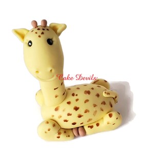 Puede incluir: Un adorno para pastel de jirafa de fondant amarillo y marr&oacute;n con manchas marrones. La jirafa est&aacute; sentada con las patas metidas debajo del cuerpo. La jirafa tiene la nariz y las orejas rosas.