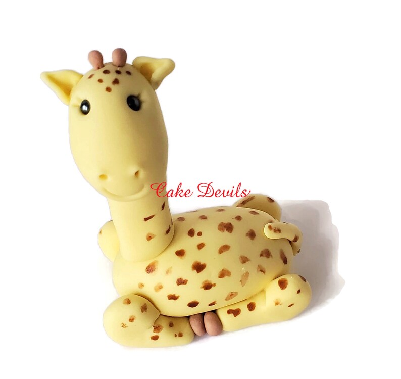 Puede incluir: Un adorno para pastel de jirafa de fondant amarillo y marr&oacute;n con manchas marrones. La jirafa est&aacute; sentada con las patas metidas debajo del cuerpo. La jirafa tiene la nariz y las orejas rosas.