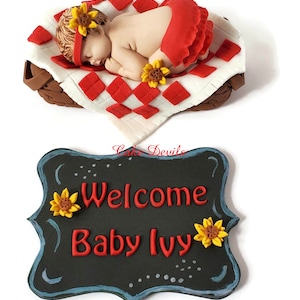May include: A red and white checked baby blanket with a sleeping baby doll on top. The baby doll is wearing a red dress and a sunflower headband. The blanket is in a brown wicker basket. A black chalkboard sign with white lettering says "Welcome Baby Ivy" with sunflowers on either side.