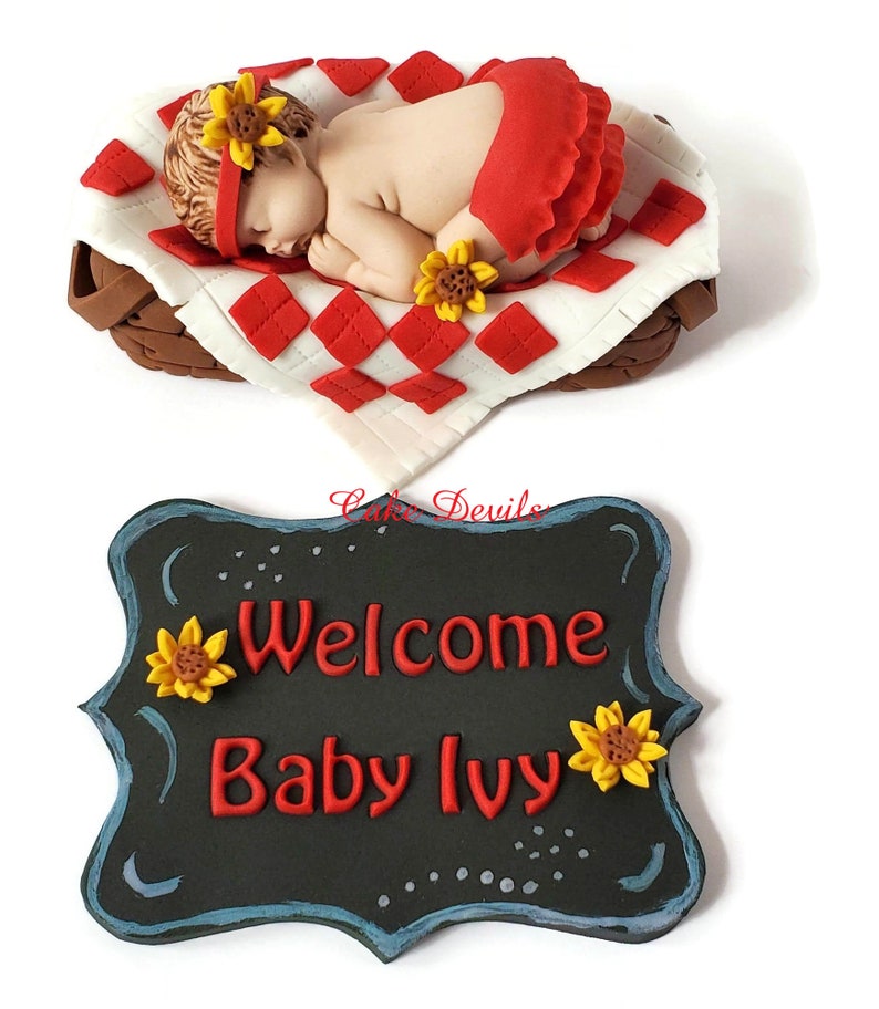 May include: A red and white checked baby blanket with a sleeping baby doll on top. The baby doll is wearing a red dress and a sunflower headband. The blanket is in a brown wicker basket. A black chalkboard sign with white lettering says "Welcome Baby Ivy" with sunflowers on either side.