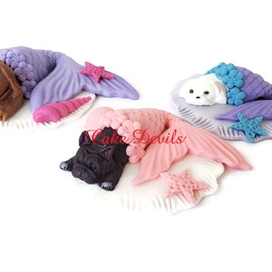 May include: Three fondant figurines of dogs dressed as mermaids. Each dog is lying on a white fondant seashell with a starfish. The dogs are a brown labrador, a white dog, and a black french bulldog. The mermaids tails are purple, blue, and pink.