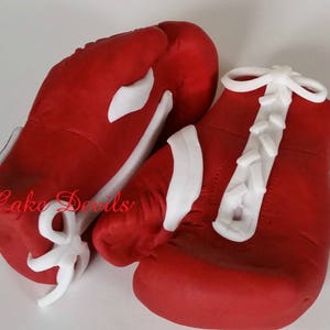 May include: Two red boxing gloves with white laces and white accents. The gloves are made of fondant and are decorated with the words "Cake Devils".