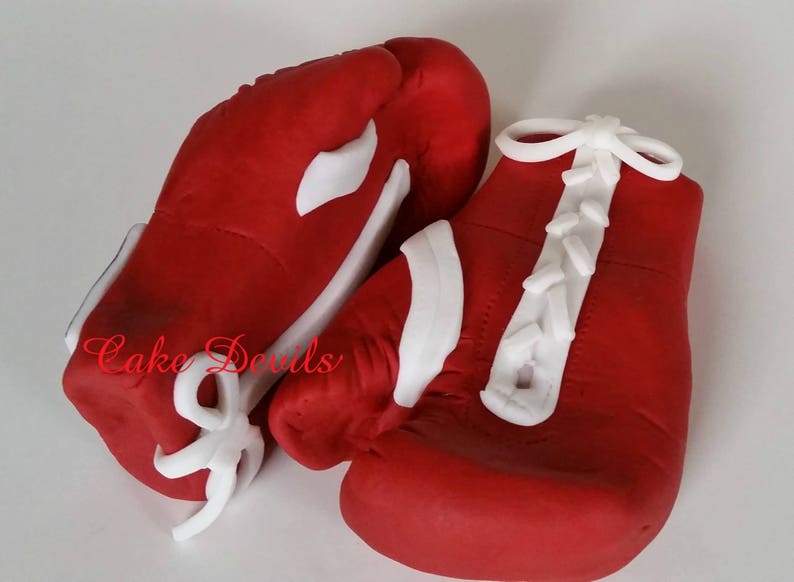 May include: Two red boxing gloves with white laces and white accents. The gloves are made of fondant and are decorated with the words "Cake Devils".