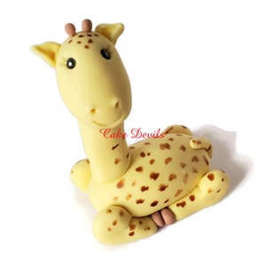 Puede incluir: Un adorno de pastel de fondant de jirafa amarillo con manchas marrones. La jirafa est&aacute; sentada con la cabeza girada hacia un lado y la boca ligeramente abierta.