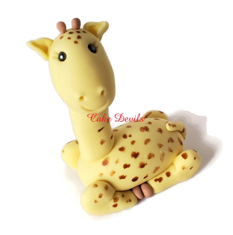 Puede incluir: Un adorno de pastel de fondant de jirafa amarillo con manchas marrones. La jirafa est&aacute; sentada con la cabeza girada hacia un lado y la boca ligeramente abierta.