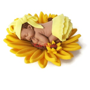 May include: A brown baby figurine wearing a yellow headband and diaper, resting on a large yellow sunflower. The sunflower has multiple layers of petals and a brown center. The figurine is a cake decoration.