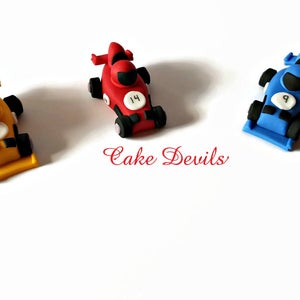 May include: Three miniature race cars made of fondant, decorated with racing numbers. The cars are yellow, red, and blue.