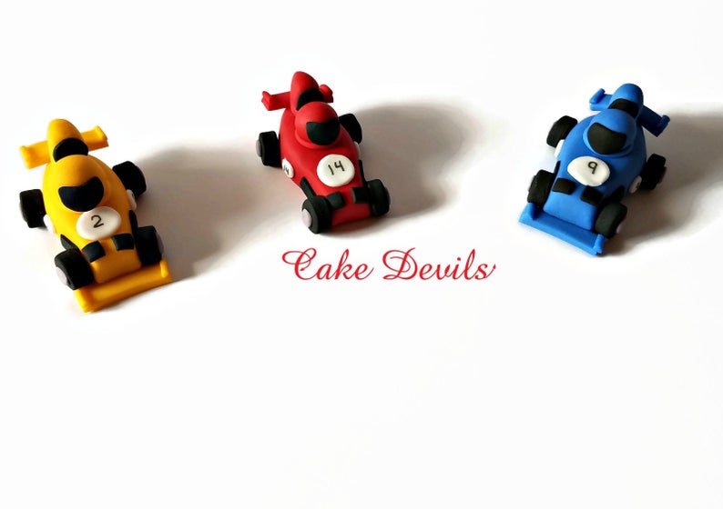 May include: Three miniature race cars made of fondant, decorated with racing numbers. The cars are yellow, red, and blue.