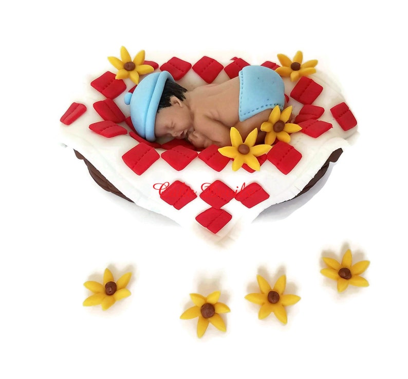 May include: A fondant baby figurine sleeping on a red and white checked blanket with yellow flower accents.
