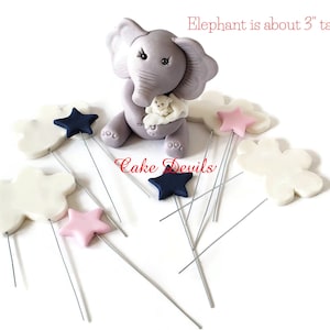 May include: A gray elephant cake topper holding a small white elephant figurine. The elephant is about 3 inches tall and is surrounded by white clouds and pink and blue stars. The stars and clouds are attached to silver wires.