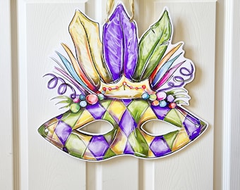 May include: A decorative Mardi Gras mask with a harlequin pattern in green, purple, and yellow. The mask features a crown and colorful feathers in yellow, purple, and green. A gold ribbon is attached for hanging.