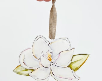 Może przedstawiać: Biały kwiat magnolii z zielonymi liśćmi, namalowany w stylu akwarelowym, jest przymocowany do brązowej wstążki z tkaniny. To ozdobny ornament.