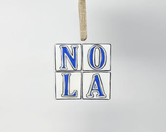 May include: A square ornament with the letters "NOLA" in blue, outlined in black, on a white background. The ornament is made of four individual squares and is suspended by a tan ribbon.