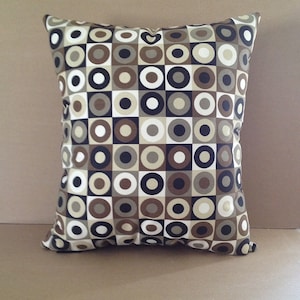 May include: Square decorative pillow with a geometric pattern of brown, tan, black, and white circles and squares. The pillow is a square shape and appears to be made of fabric. The design is a repeating pattern.