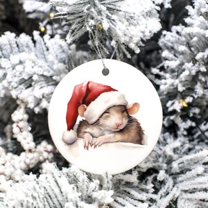 Peut inclure: Une boule de Noël ronde en céramique blanche avec une illustration aquarelle d'un rat brun portant un bonnet de Père Noël rouge. Le rat dort les yeux fermés et ses pattes sont repliées sous son menton.