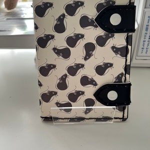 May include: A white fabric notebook cover with a black and white mouse pattern. The cover has two black leather straps with a white button on each.