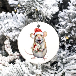 May include: A circular Christmas decoration depicting a smiling mouse in a Santa hat, clutching a bouquet of red and white flowers. The ornament has a white backdrop and a silver hanging string. The mouse has tan and white fur.