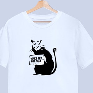Pode incluir: Camiseta branca com um gráfico preto e branco de um rato segurando um cartaz que diz "Make Tea Not War."