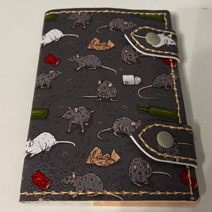 May include: A dark grey leather wallet featuring a pattern of grey and white rats, with scattered rubbish and bottles. The wallet has a snap closure and gold stitching around the edges.