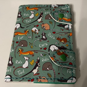 Può includere: Un portafoglio in pelle verde con un motivo a ratti. Il portafoglio ha una patta con chiusura a scatto e una piccola tasca sul davanti. Il portafoglio è decorato con diversi ratti in pose diverse, tra cui alcuni con il formaggio. Il testo "Ratti", "Coda", "Pelliccia", "Orecchie", "Denti" e "Piedi" è stampato sul portafoglio.