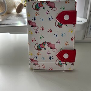 May include: A white leather wallet with a colorful rat print and red accents. The rats are wearing colorful hats and holding paintbrushes. The wallet has two red straps with white buttons.