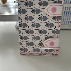 May include: A white leather card holder with a pink stitched border and a repeating pattern of gray mice. The card holder has a white flap with a pink button and a pink stitched border.