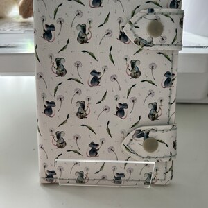 May include: A white wallet with a repeating pattern of watercolor mice holding dandelions. The wallet has a snap closure and a clear plastic stand. The design includes green leaves and dandelion seed heads.
