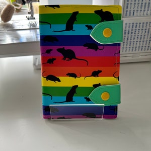 May include: A rainbow-colored wallet with a green flap and a yellow button. The wallet is decorated with a pattern of black rats.