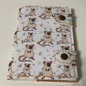 May include: A white leather notebook cover with a repeating pattern of brown mice and dandelion seeds. The cover has two brown leather flaps with gold stitching and a gold button closure.