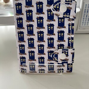 May include: A white leather wallet with a repeating pattern of blue police boxes and brown rats. The wallet has two white snap closures.