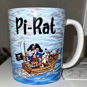 May include: A white ceramic mug with a blue and white illustration of a rat pirate and a dog in a boat. The text "Pi-Rat" is printed in black on the mug.