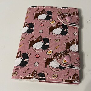 May include: A pink leather wallet with a repeating pattern of black and brown rats with white bellies. The rats are clustered together and appear to be cuddling. The wallet has two pink snaps on the right side.