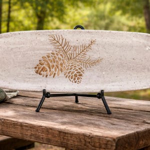May include: A rectangular, speckled ceramic serving platter with a pine cone design in shades of brown. The platter is displayed on a black metal stand, set on a wooden table. A green and white plaid cloth is partially visible.