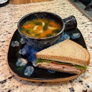 May include: A bowl of chicken vegetable soup with carrots and greens served alongside a turkey, cheese, and lettuce sandwich on whole wheat bread. The soup and sandwich are presented on a dark blue and black speckled ceramic plate and bowl set.
