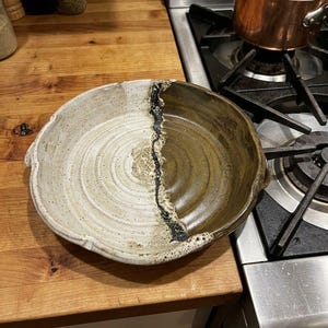 May include: A round ceramic bowl featuring a split design with one half in a speckled off-white glaze and the other half in a dark olive green glaze, separated by a textured, dark, and uneven vertical line running through the center.