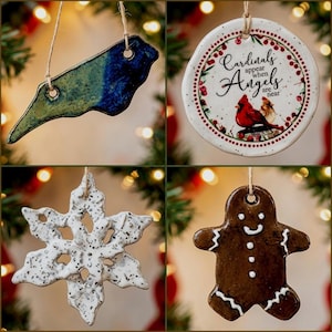 May include: Four ceramic ornaments hanging by twine. Top left: a blue and green North Carolina state shape. Top right: a round white ornament with red berries, a cardinal, and the text Cardinals appear when Angels are near. Bottom left: a white speckled snowflake. Bottom right: a brown gingerbread person.