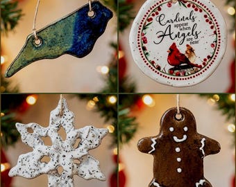 May include: Four ceramic ornaments hanging by twine. Top left: a blue and green North Carolina state shape. Top right: a round white ornament with red berries, a cardinal, and the text Cardinals appear when Angels are near. Bottom left: a white speckled snowflake. Bottom right: a brown gingerbread person.