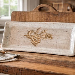 May include: A rectangular, off-white ceramic serving tray with a gold pinecone design, resting on a wooden cutting board. A small cheese knife and a folded linen napkin are in the foreground. The cutting board has a handle.