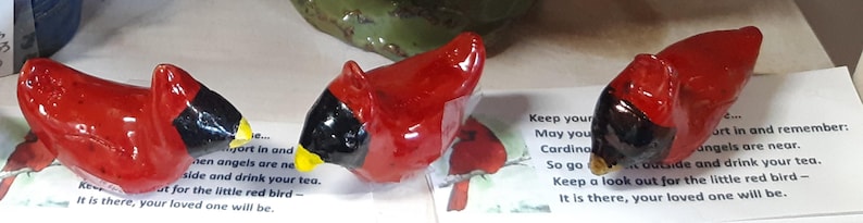 May include: Three small ceramic cardinal figurines with black and red paint. The figurines are sitting on a white surface with a card that reads "Keep your heart in and remember: angels are near. Go outside and drink your tea. Keep a look out for the little red bird - It is there, your loved one will be."