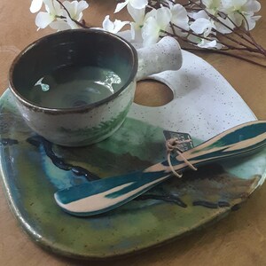 May include: A white ceramic serving dish with a green and blue glaze. The dish has a small, round indentation in the center and a small, white ceramic bowl with a green and brown glaze. A wooden spoon with a green and white glaze is on the dish.