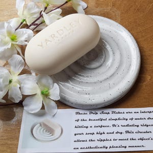 May include: White ceramic soap dish with a rippled design. The dish is designed to hold a bar of soap and allow water to drain. A bar of Yardley London soap is sitting on the dish.