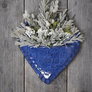 May include: A heart-shaped blue ceramic wall pocket planter with a scalloped edge and embossed house design, filled with a bouquet of artificial light green foliage and small white flowers, creating a decorative wall-mounted display.