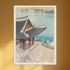 May include: An Asian-inspired art print featuring a traditional pagoda with a dark gray roof and red accents, overlooking a serene blue lake. Cherry blossoms in full bloom frame the foreground, with figures in traditional attire. The artwork is framed in white.