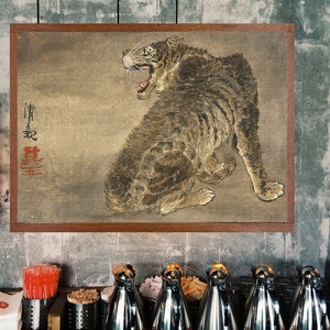 K&ouml;nnte beinhalten: Ein gerahmtes Gem&auml;lde eines braunen und wei&szlig;en Tigers mit gr&uuml;nen Augen, der mit offenem Maul br&uuml;llt. Das Gem&auml;lde ist auf einem beigefarbenen Hintergrund mit roten japanischen Schriftzeichen. Das Gem&auml;lde h&auml;ngt an einer grauen Wand.