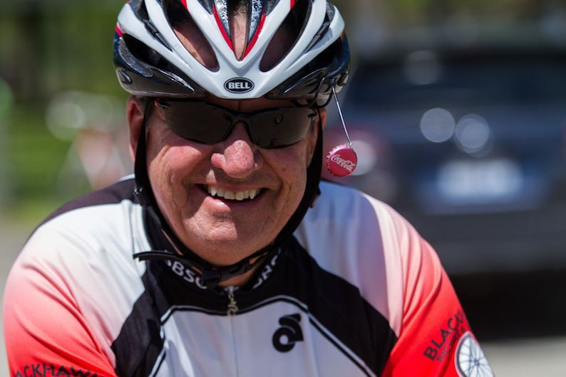 May include: A man wearing a white, black, and red bicycle helmet with a black visor and sunglasses. He is smiling and wearing a white and black cycling jersey with red accents. A red Coca-Cola bottle cap is hanging from his helmet.