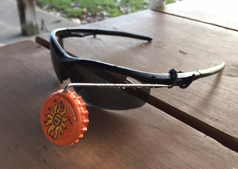 May include: Black sunglasses with a silver metal frame and a black rubber strap. A bright orange bottle cap with a sun design is attached to the frame.