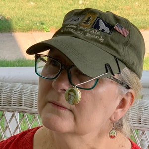 May include: A woman wearing a green baseball cap with the text "Isle Royale National Park" and an American flag patch. She is also wearing glasses and a cherry-themed earring.