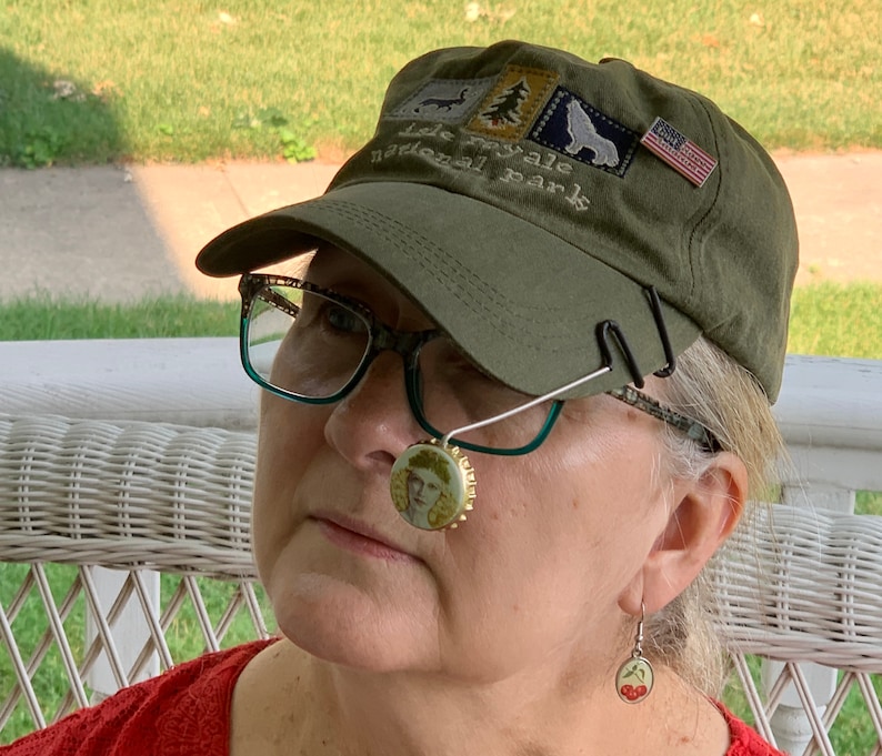 May include: A woman wearing a green baseball cap with the text "Isle Royale National Park" and an American flag patch. She is also wearing glasses and a cherry-themed earring.